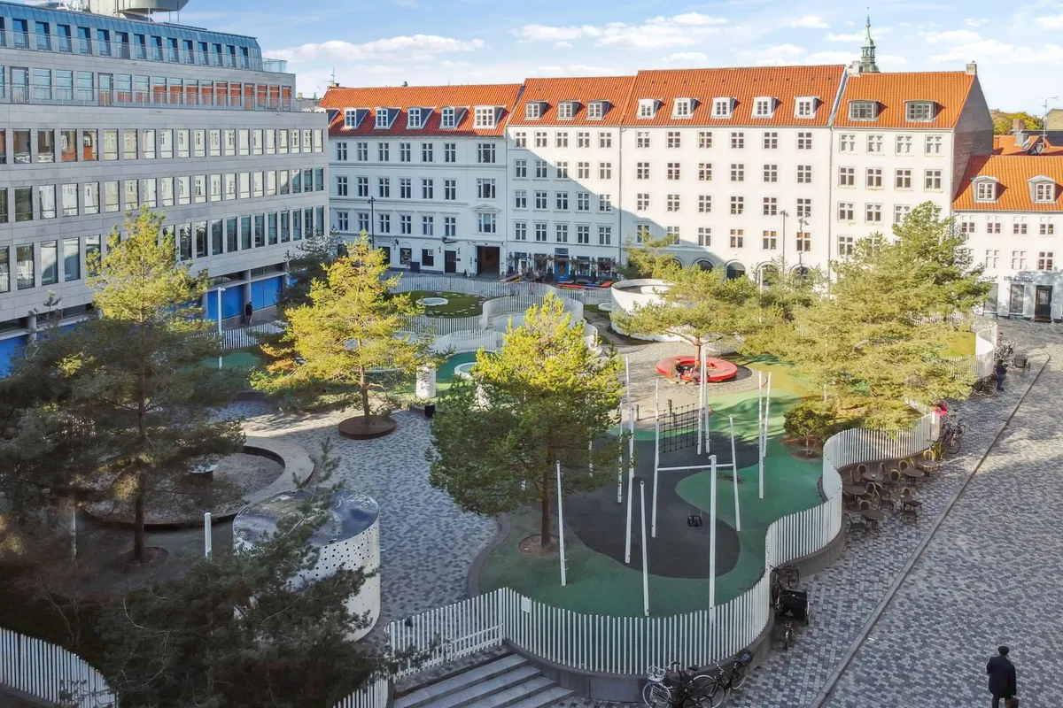 Åben plads med træer, grønne områder og legeplads. Moderne bygninger med røde tag. Gade med søjler, parkerede cykler og