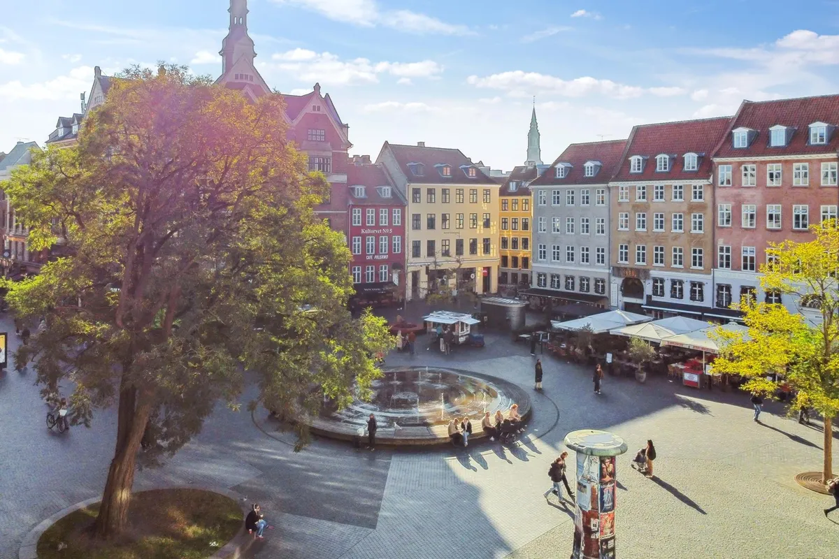 Mennesker går og sætter sig på Hauser Plads i København, med en kilde og forskellige bygninger og træer i baggrunden