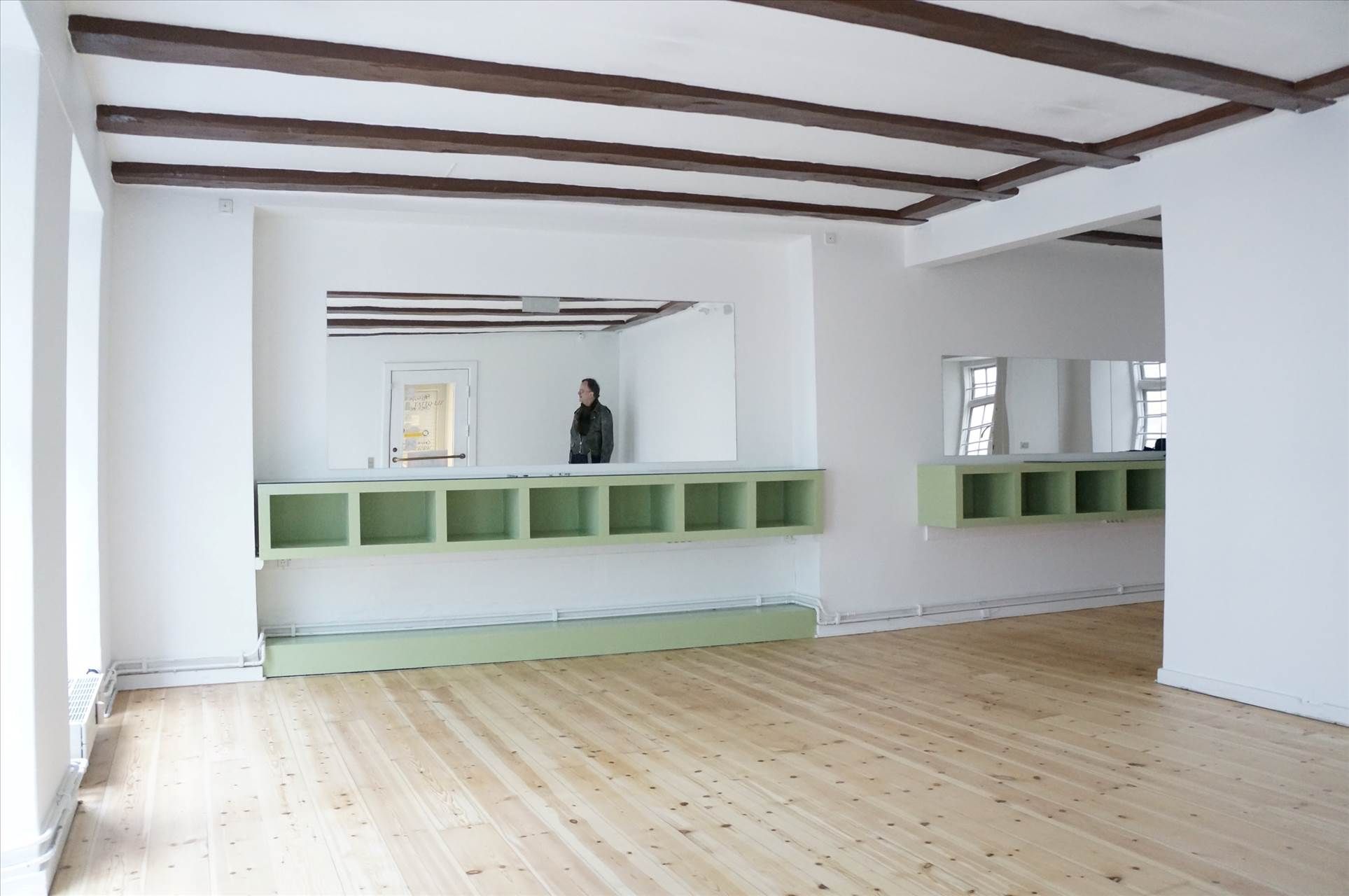 En kvinde står foran et spejl i en fredede bygning. Væggene er hvide og loftet er træ. Der er et grønt hyldesystem og gulvet