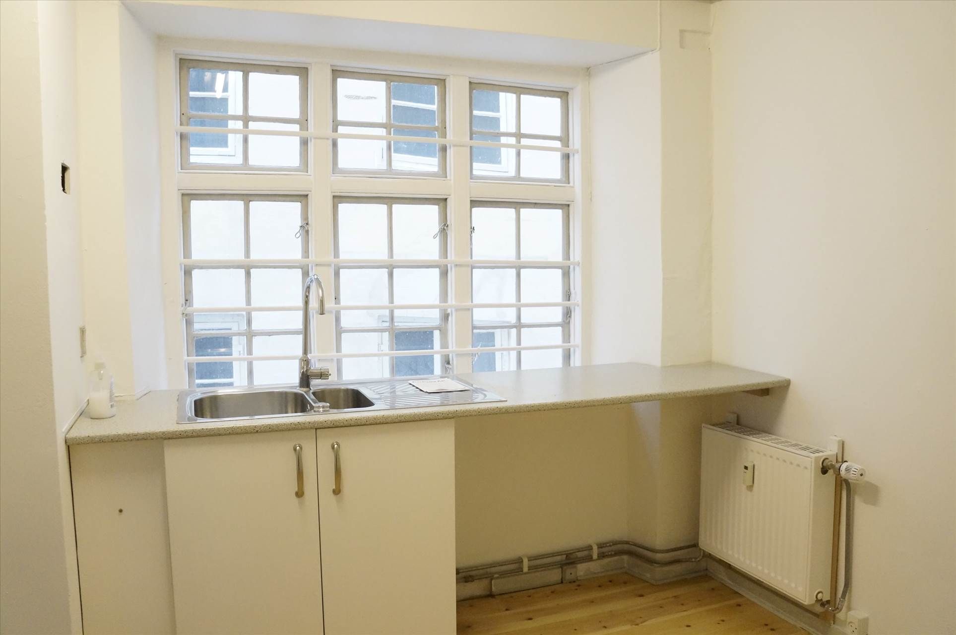 Lyset skinner ind i et lille køkken med vindue og radiator. Væggene er hvide og der er et nyt gulv.
