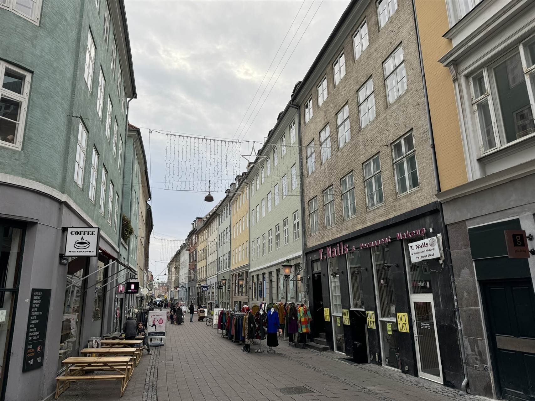 Butikker med klæder og mad på en gågade i København. To borde med sæder udenfor og en person læser på plakat.