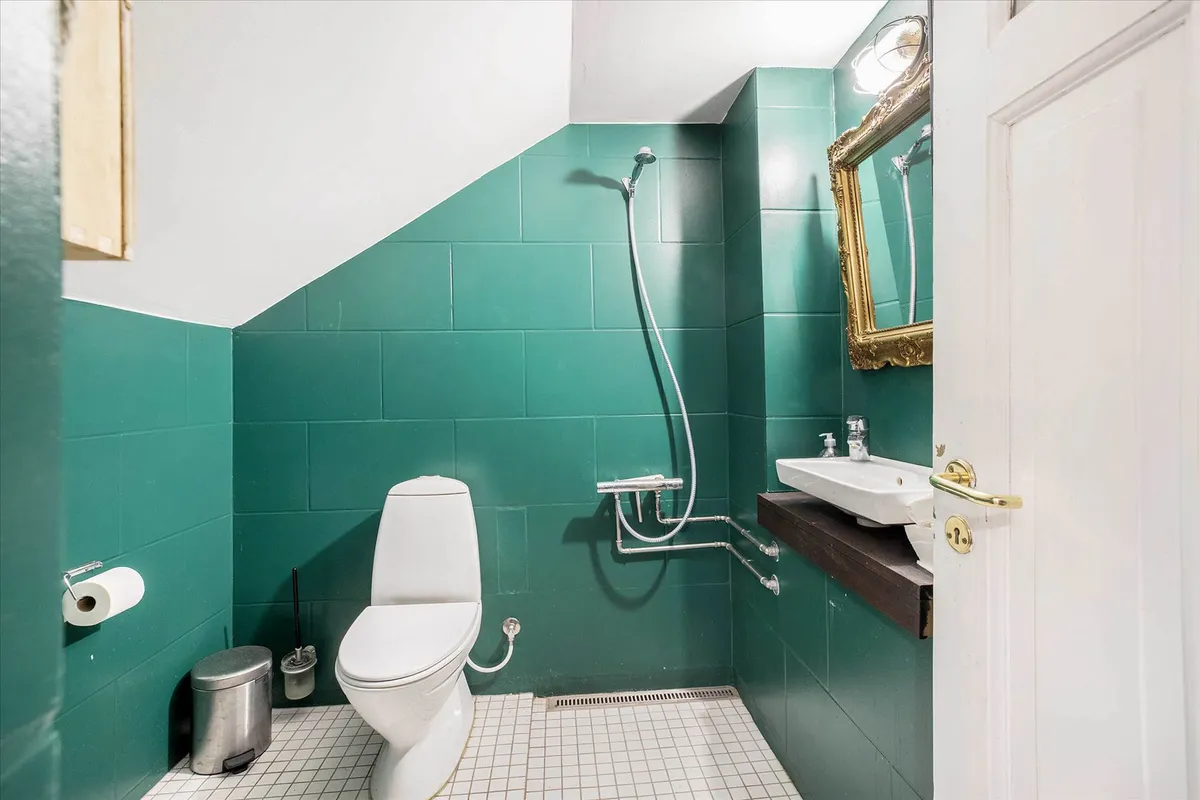 Det grønne badeværelse med hvidt toilet, håndvask og grønne vægge har en åben dør med guldbeslag