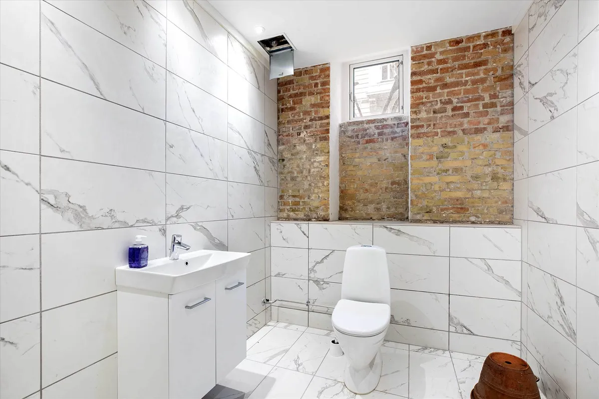 Et lille badeværelse med en hvid toilet, hvid vask og marmorblegede vægge og gulv samt en rødmalet krukke i hjørnet