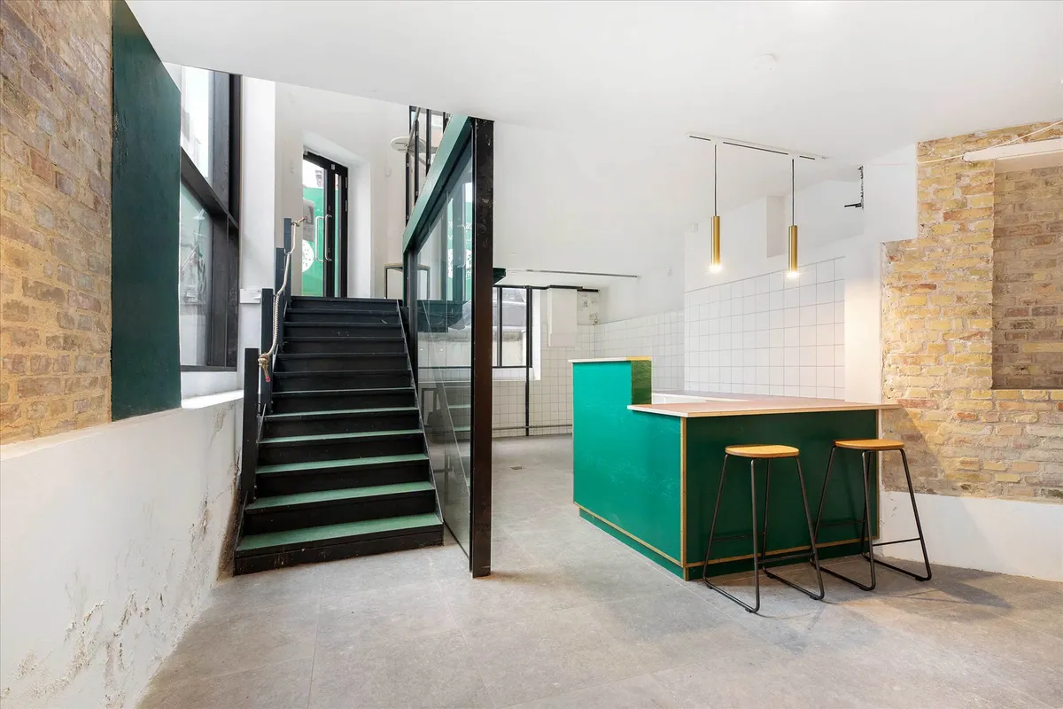 Inde i et hus er der et åbent køkken med grønne stole og en grøn væg. Stairs er op til næste etage. Loftet har lysekroner,