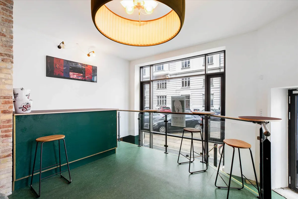 Inde i en elegant indretning med gulvtæppe og loftslampe er der en grøn bardisk med tre høje stole og en vase med solmotiv