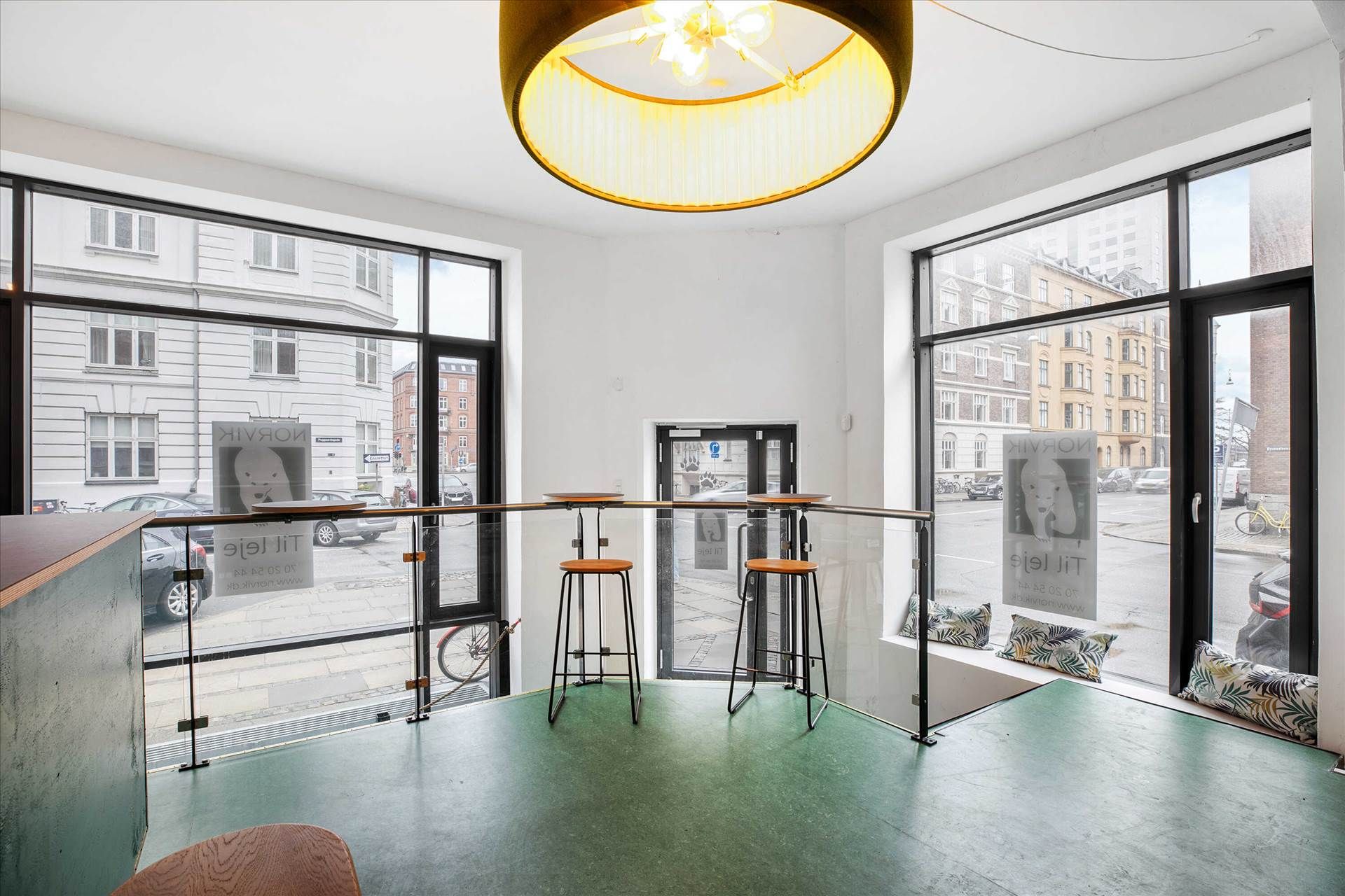 Vinduesfront af lokale med 2 høje, lyse barstole og lysegul loftslampe i et område med biler, cykler og grønne områder