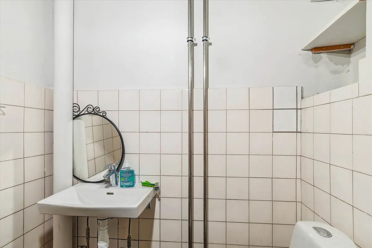 Et lille badeværelse med en hvid mur og en hvid toilet i højre side. En hvid vask med et spejl og håndvask samt en dispenser