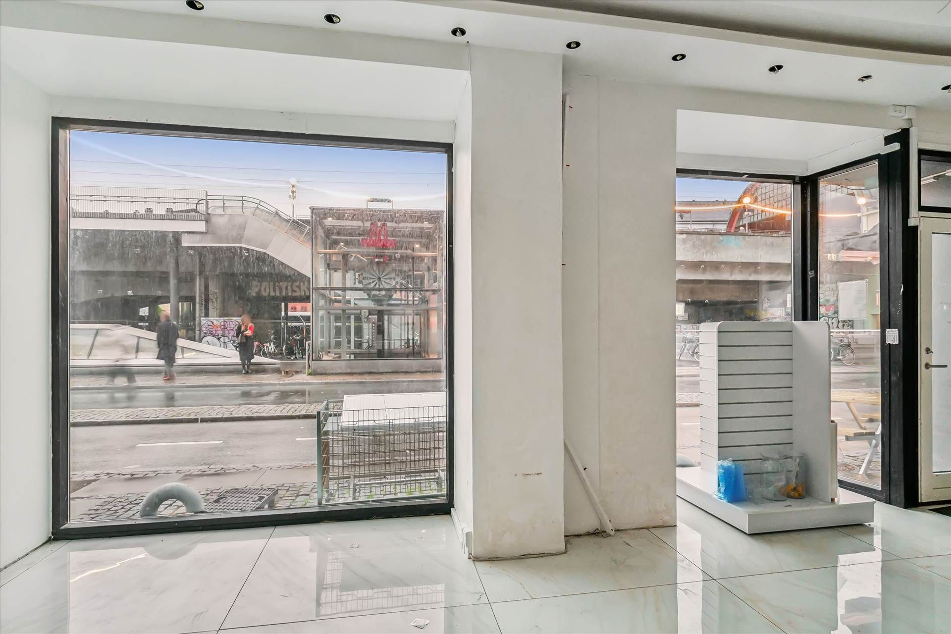 Et tomt lokale med store ruder og hvide vægge og gulv samt et højt loft. Udsigten er til en gågade med overgange og nogle