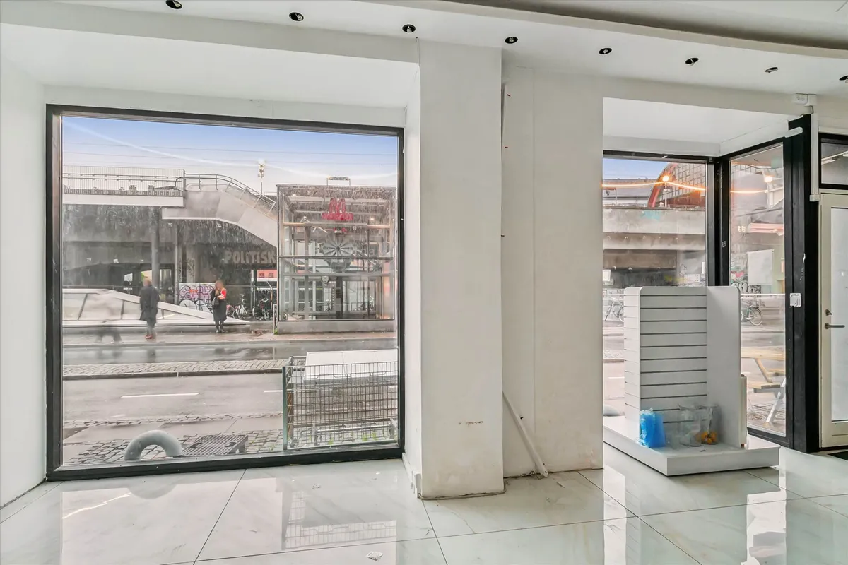 Et tomt lokale med store ruder og hvide vægge og gulv samt et højt loft. Udsigten er til en gågade med overgange og nogle