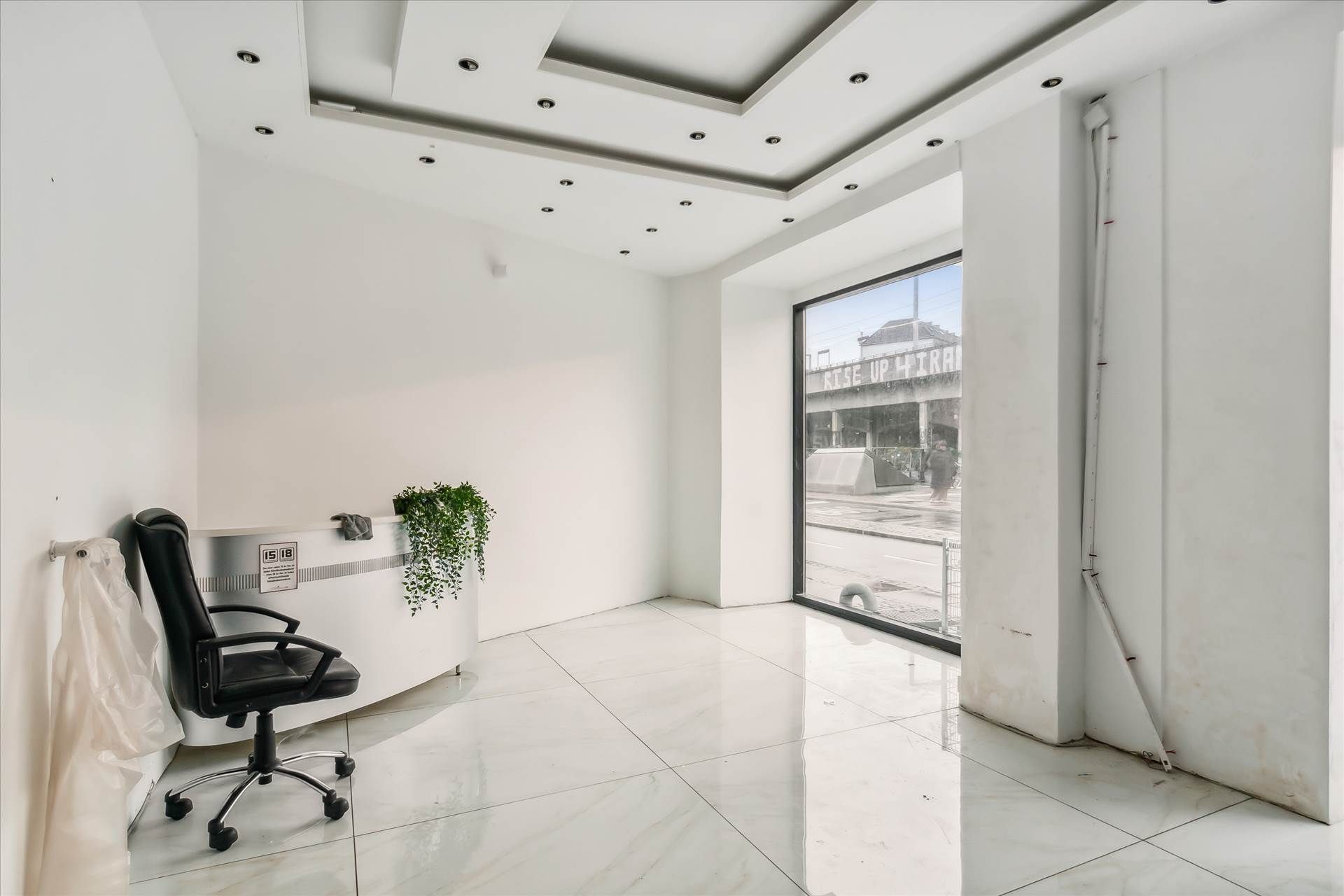 En tom hvid stue med lysegulve, højt til loftet og en stor glasdør, der fører til en travl gade.