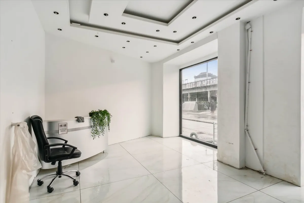 En tom hvid stue med lysegulve, højt til loftet og en stor glasdør, der fører til en travl gade.