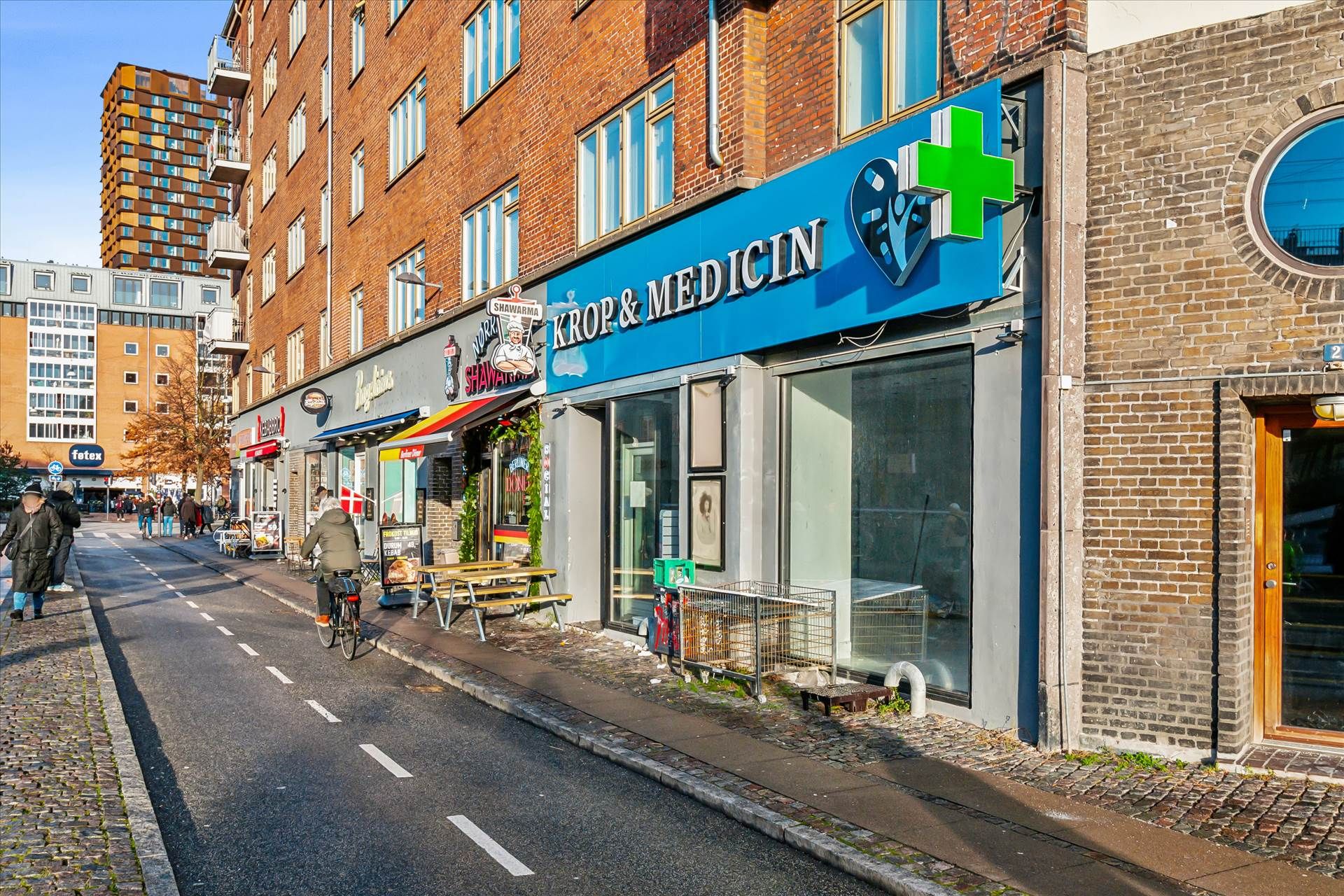 Mennesker går på fortovet foran et apotek og et café med udendørs spisning. Et enkeltcykel cykler på en cykelstig, og der er
