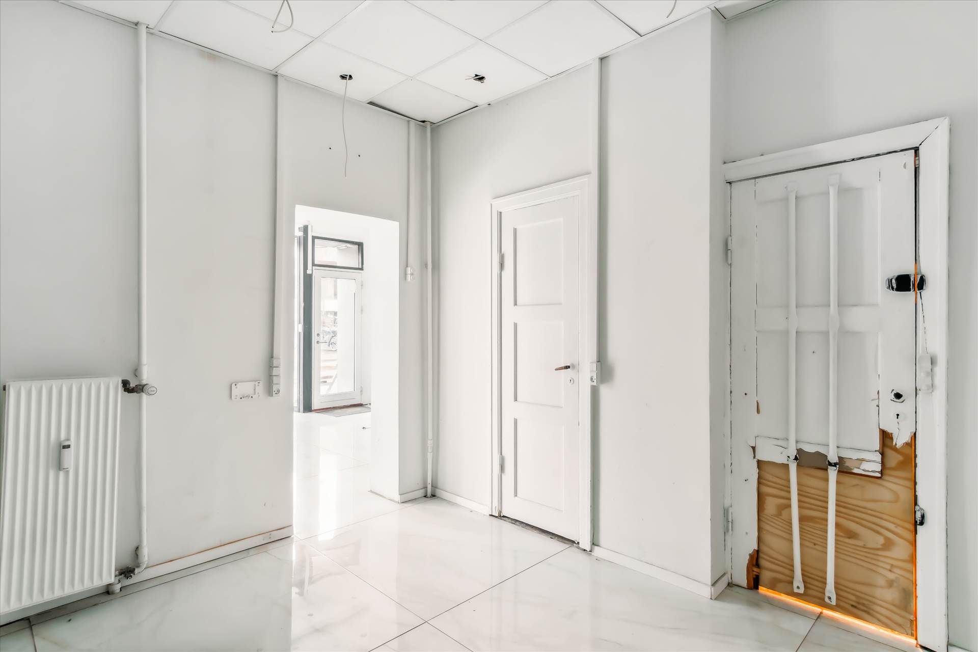 Ledig 69 m² butik med hvide vægge og moderne indretning og gulvbelægning, to døre og radiator samt lyse vinduer