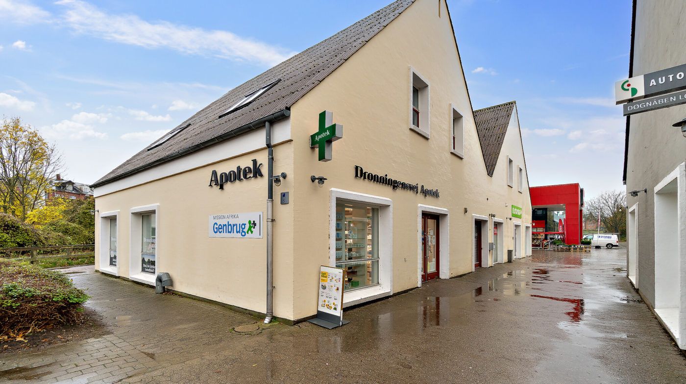 Apotek med Genbrug og Dronningensvej Apotek skiltet. Foran apoteket er der et tomt parkeringsområde.