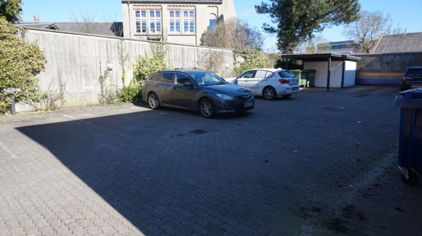 To biler parkeret i en parkeringsplads med en hvid mur og grønne affaldsspande bag ved en bygning med flere vinduer