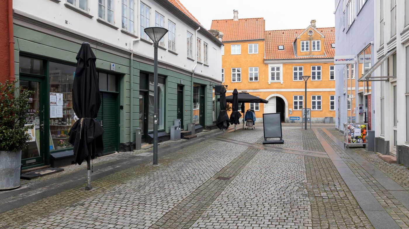 Bred gadegade med to butikker med udstående parasoller og en tom højstol og en stol med tøj i en butik i baggrunden