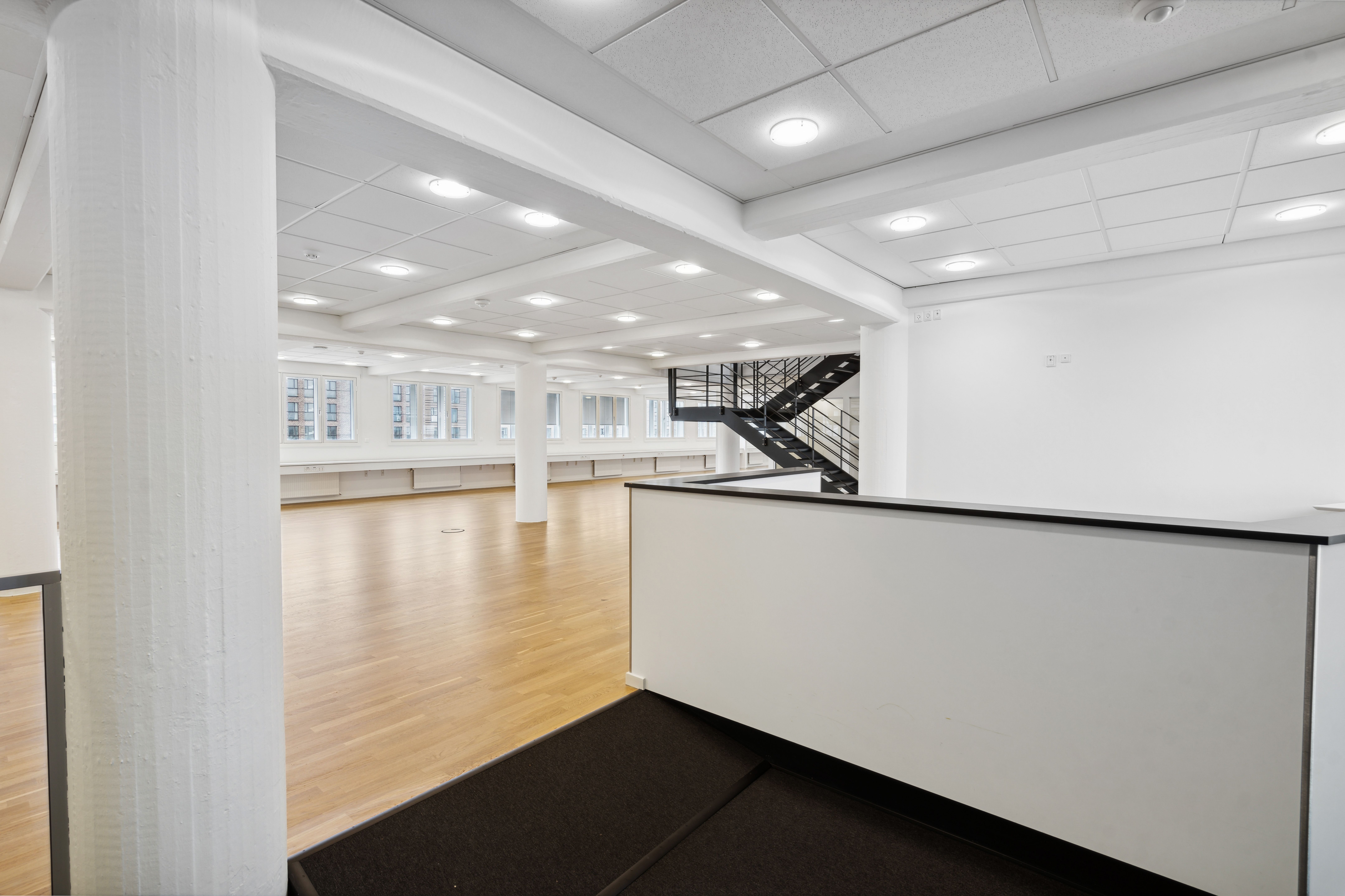 En stor, tom kontorlokal med højloft og lysegul trægulv. Lokalet har moderne hvide vægge og loftslampe. Der er en bred gang
