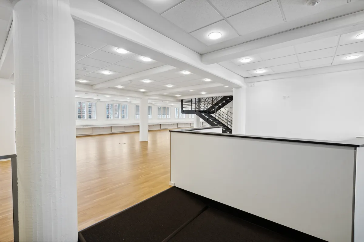 En stor, tom kontorlokal med højloft og lysegul trægulv. Lokalet har moderne hvide vægge og loftslampe. Der er en bred gang