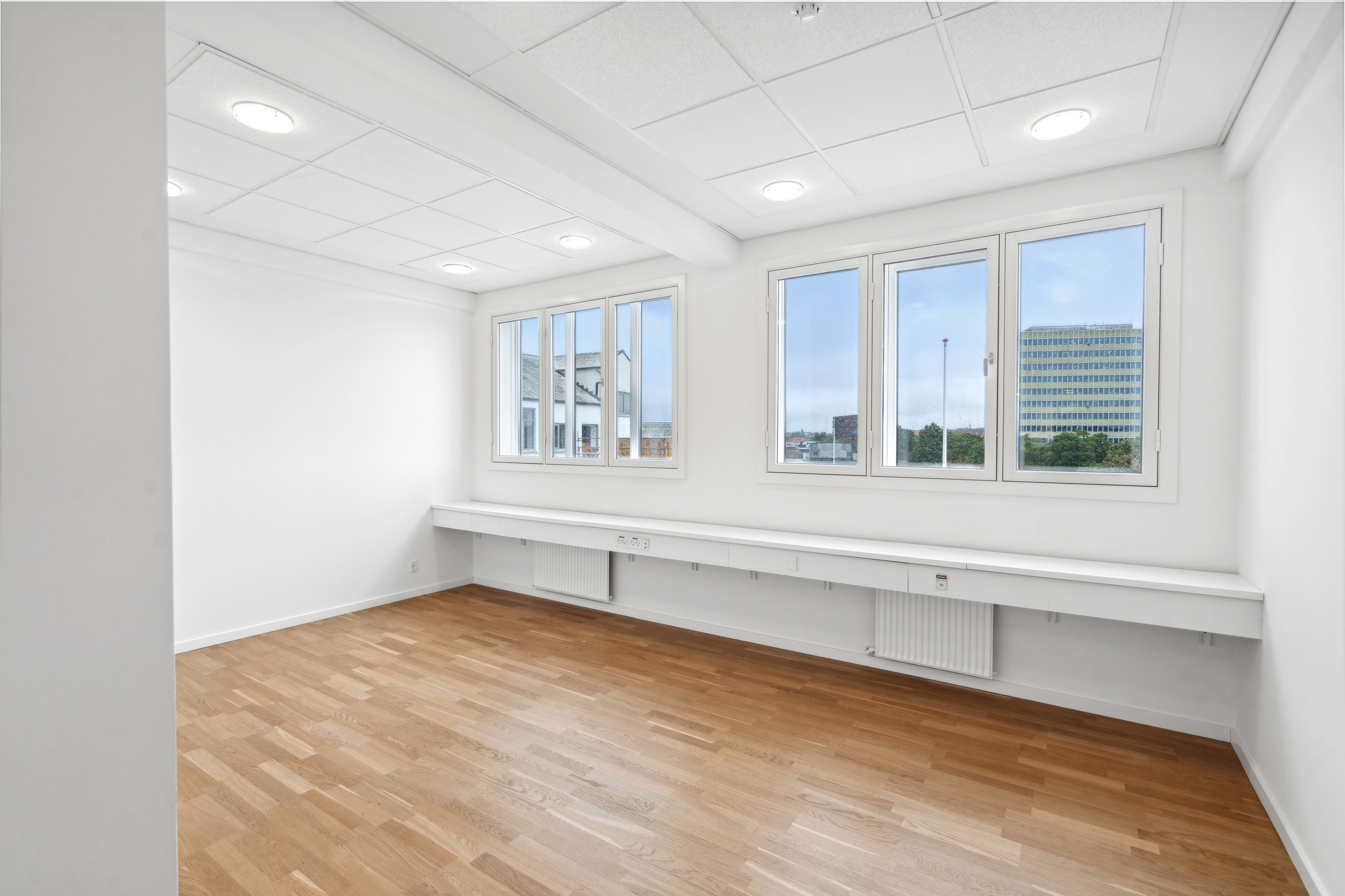 Et stort rum med hvide vægge, lyse trægulve og tre store vinduer med udsigt til et bygningskompleks og grønne træer