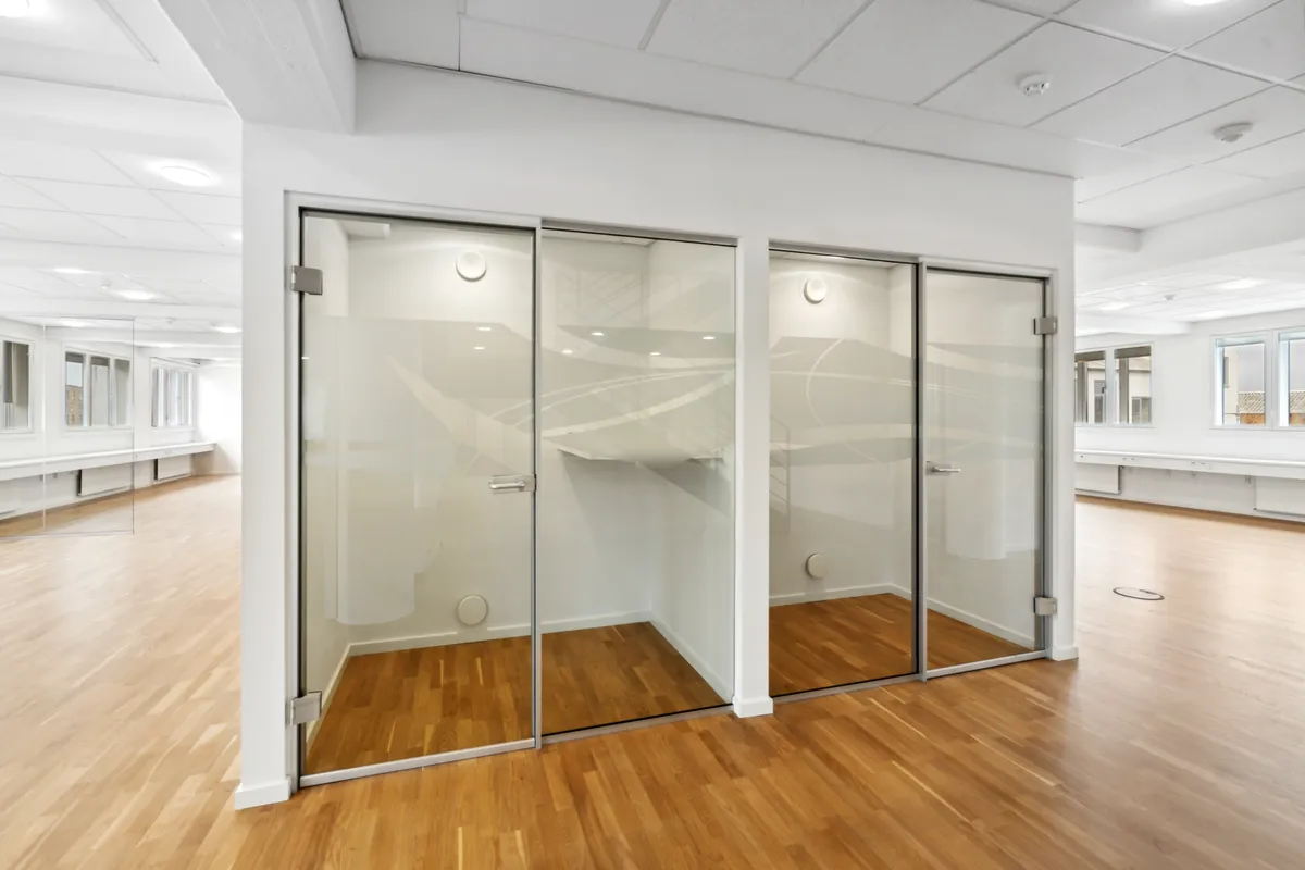 Minimalistisk kontor med glasvægge og højloft. Der er lysegule trægulve og hvide vægge. Loftet er hvidt og pænt oplyst af