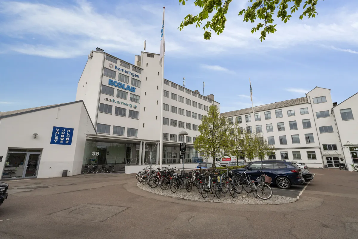Et stort hvidt bygningskompleks med flere lokaler og Ecolab-skilt, parkeringsplads med cykler og biler samt en gågade foran