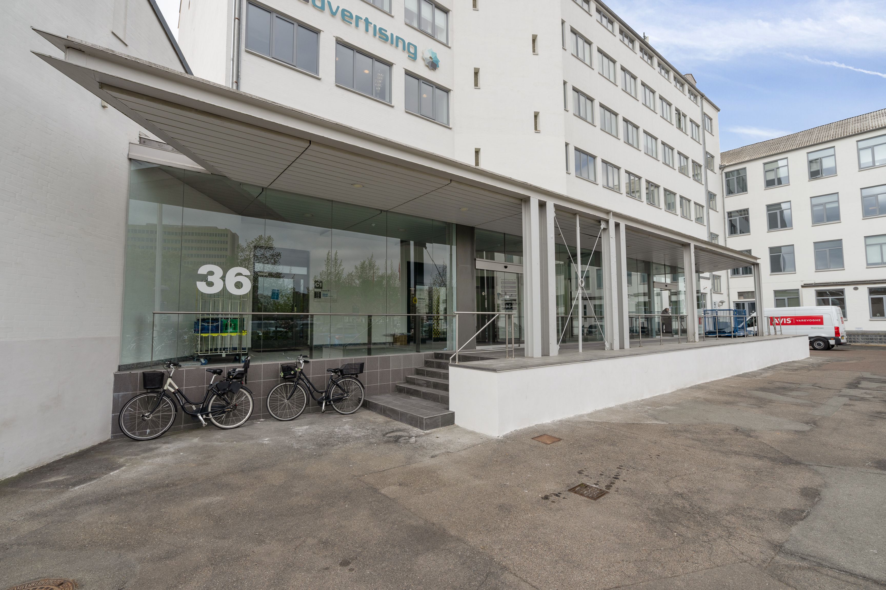 Bygningens facade har store vinduer, glasdør og et stort "36" i nummerpladen. To cykler er parkeret foran indgangen, og der