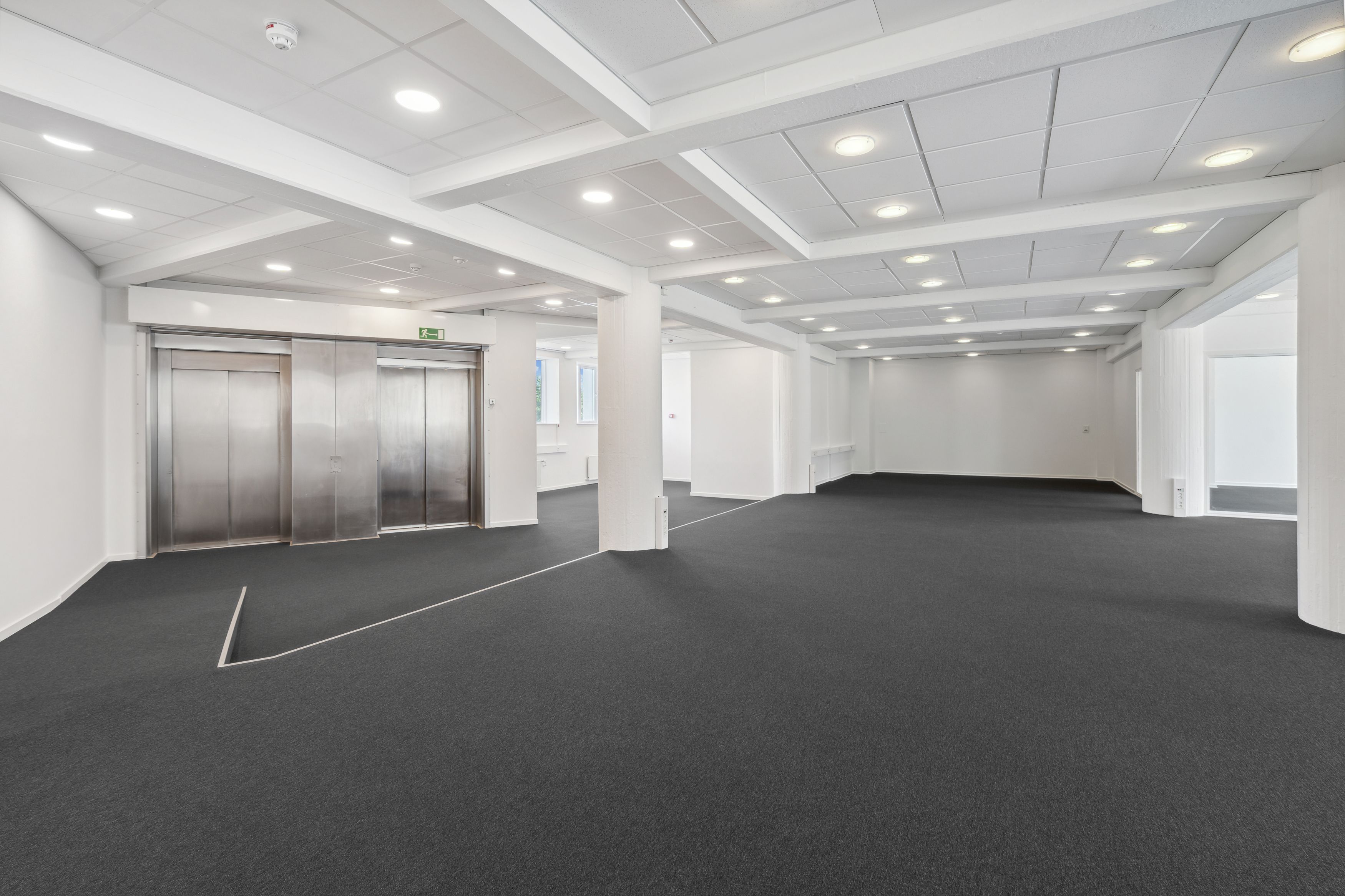 Moderne og neutralt indrettet lokale med højt til loftet og lyse loftsbjælker. To elevatorer, hvide vægge og gulve med grå