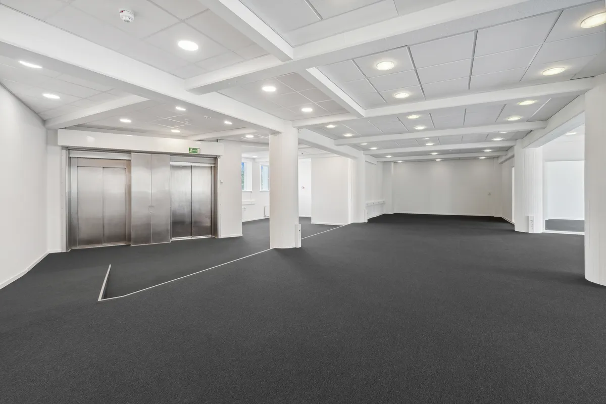 Moderne og neutralt indrettet lokale med højt til loftet og lyse loftsbjælker. To elevatorer, hvide vægge og gulve med grå