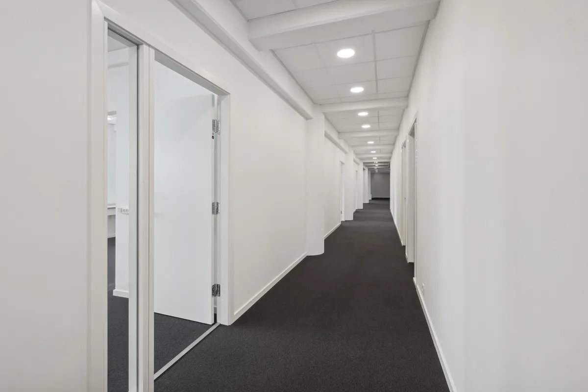 Lang hal med hvide vægge og gulv og mørkt gulvtæppe i centrum to glasdøre og hvide loftsbælte med indbyggede lysekasser