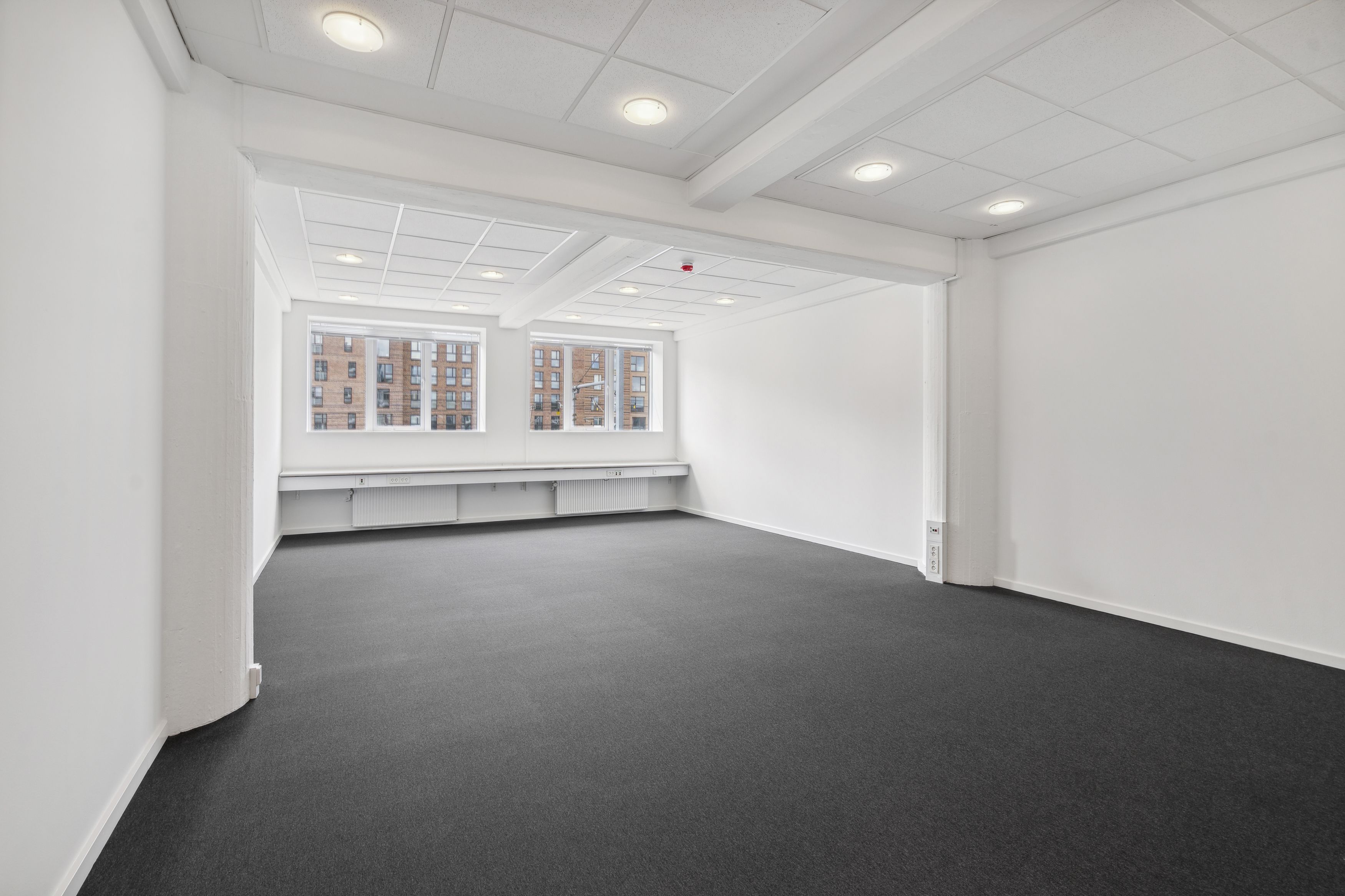 Et rum med hvide vægge og gulv, mørkt gulvtæppe og hvide loftsbelægninger med indbygget belysning. To store vinduer med