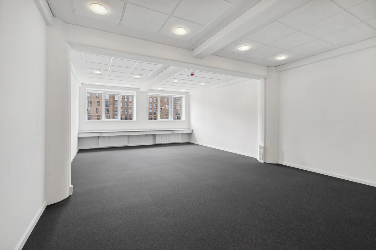 Et rum med hvide vægge og gulv, mørkt gulvtæppe og hvide loftsbelægninger med indbygget belysning. To store vinduer med