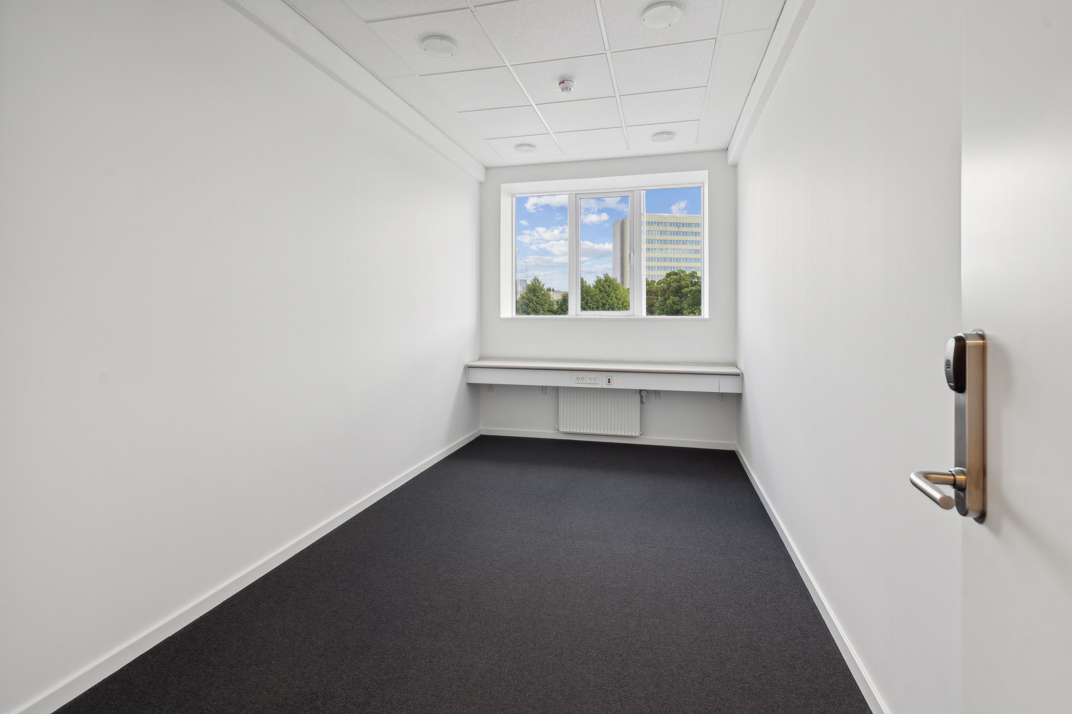 Moderne, tomt kontor med hvide vægge, gulv, loft, slanke radiator og et vindue med udsigt til bygninger og grønne træer