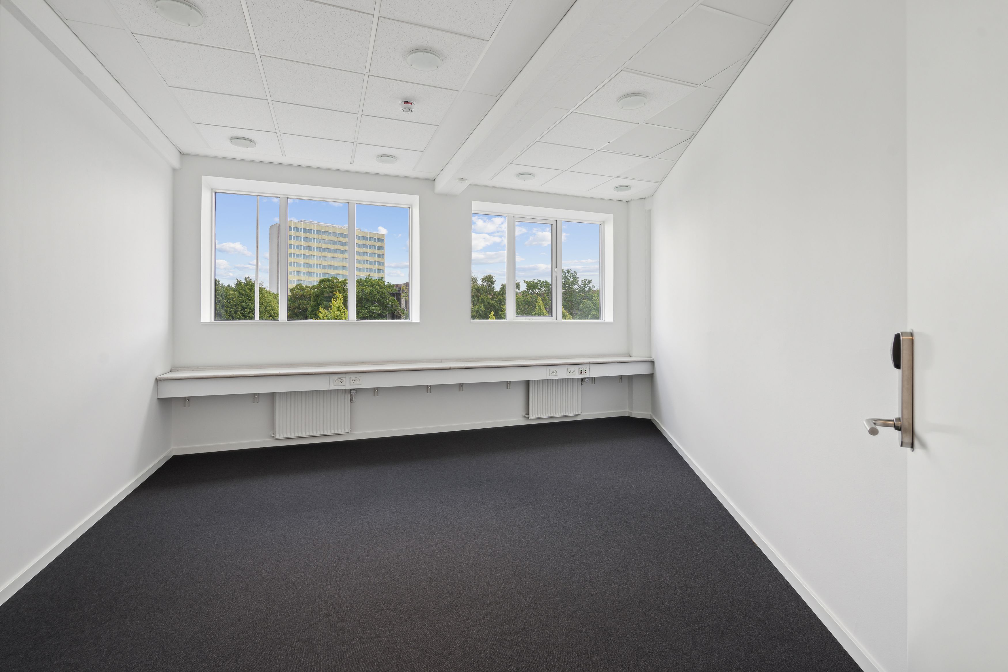 To vinduer med udsigt til et bygningskompleks og træer, hvid væg og hvid loftsbeklædning med indlejrede rundede lys, sort