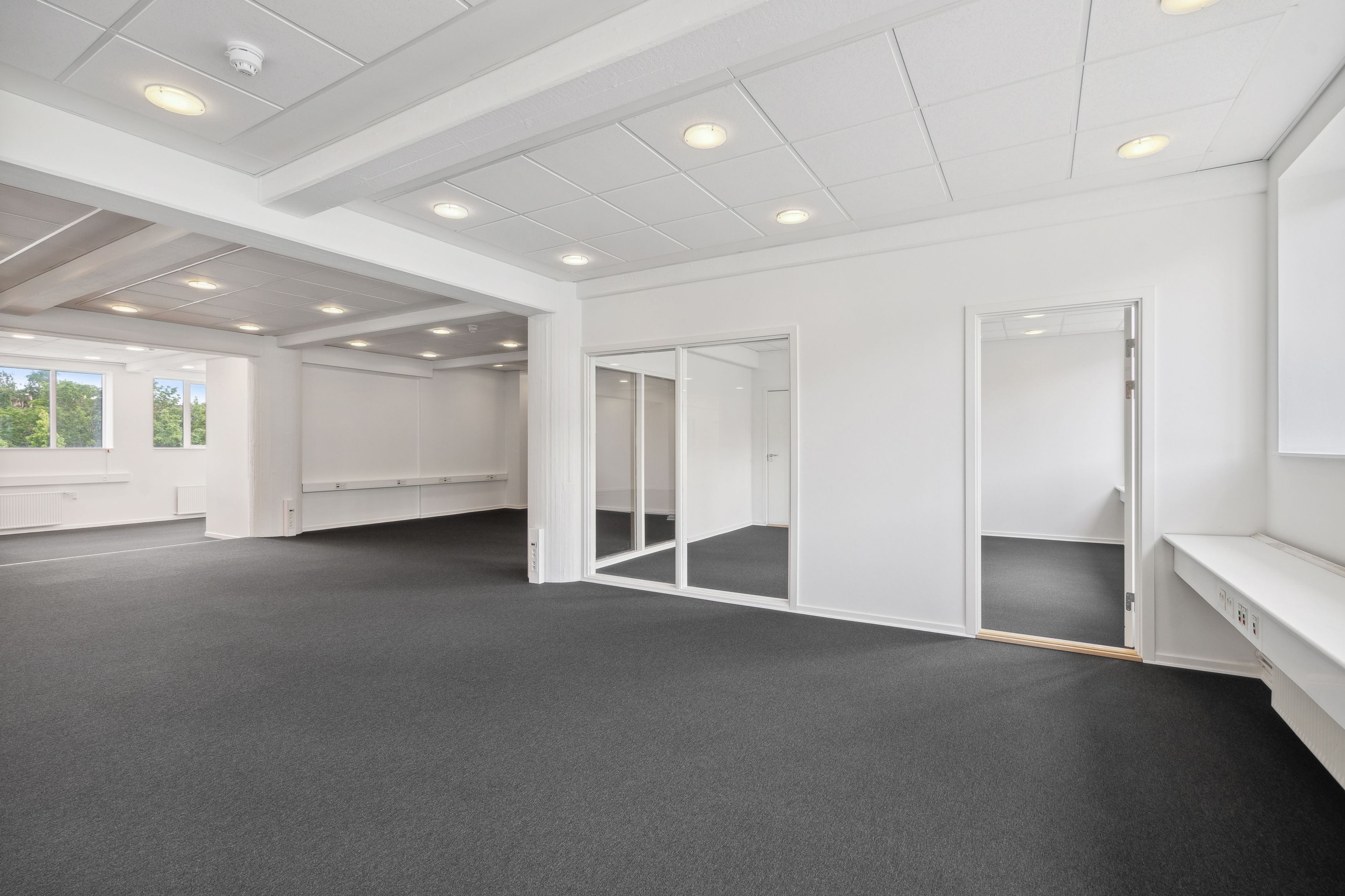 Værelse med glasdøre og hvide vægge, gulvbelægning og flere indbyggede lyspunkter på loftet, der er et vindue med udsigt til