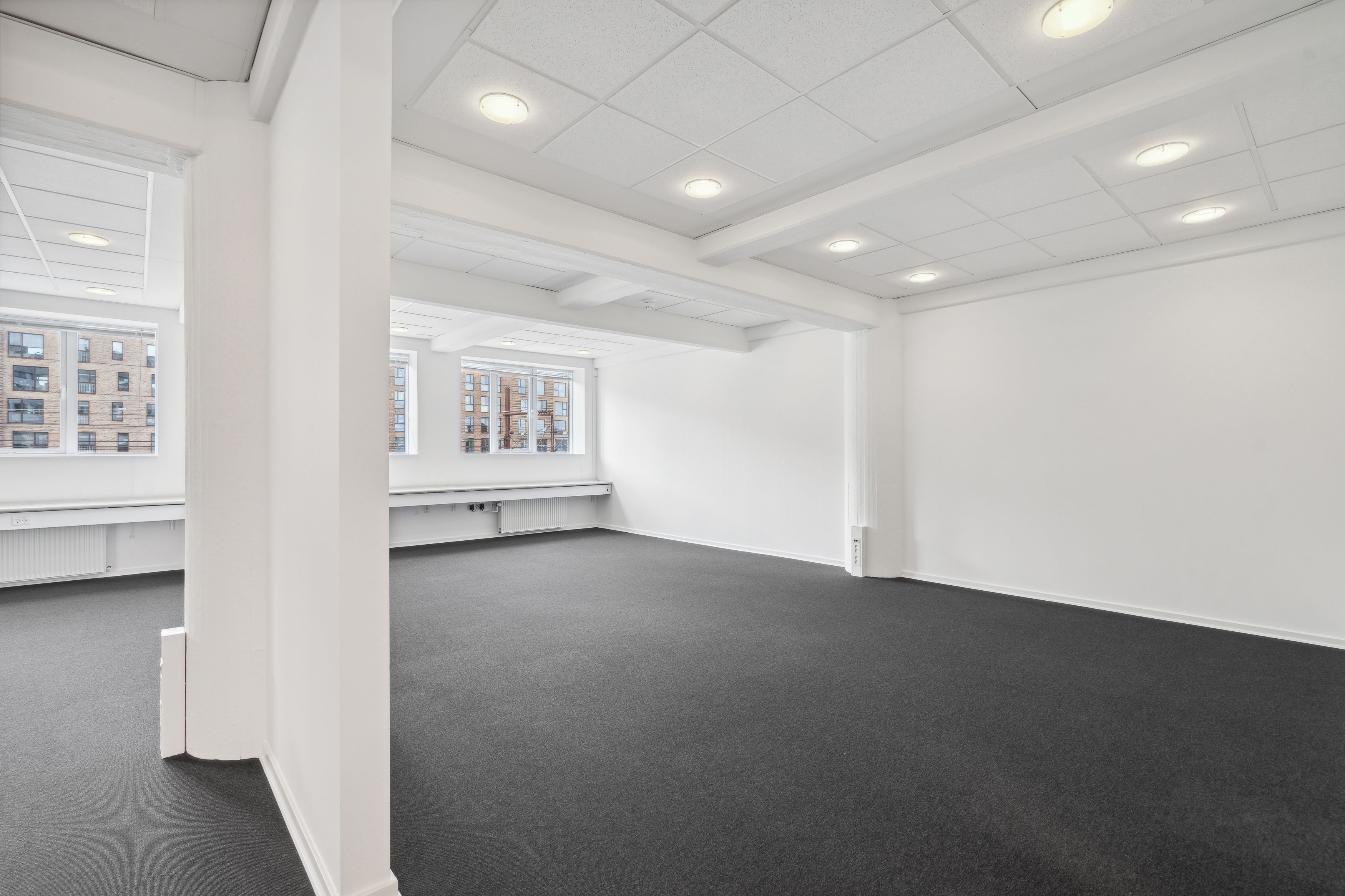 To lokaler med store vinduer, hvide vægge og mørk gulvbelægning. Værelset er tomt med flere indbyggede radiatorer og flere