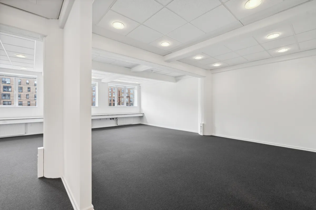 To lokaler med store vinduer, hvide vægge og mørk gulvbelægning. Værelset er tomt med flere indbyggede radiatorer og flere