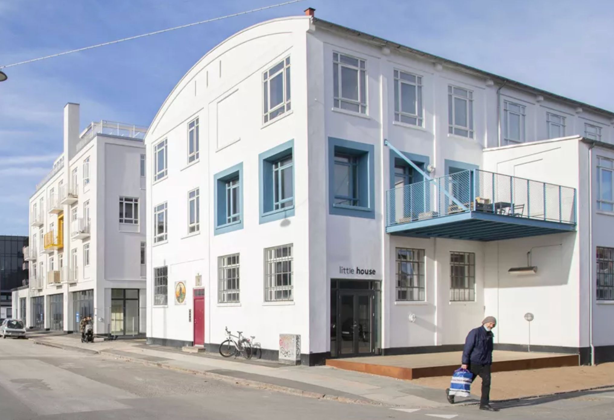 Mand med poser går forbi et hvidt bygningskompleks med blå vinduesindfatninger og rødt dørblad. På bygningen står navnet Little House