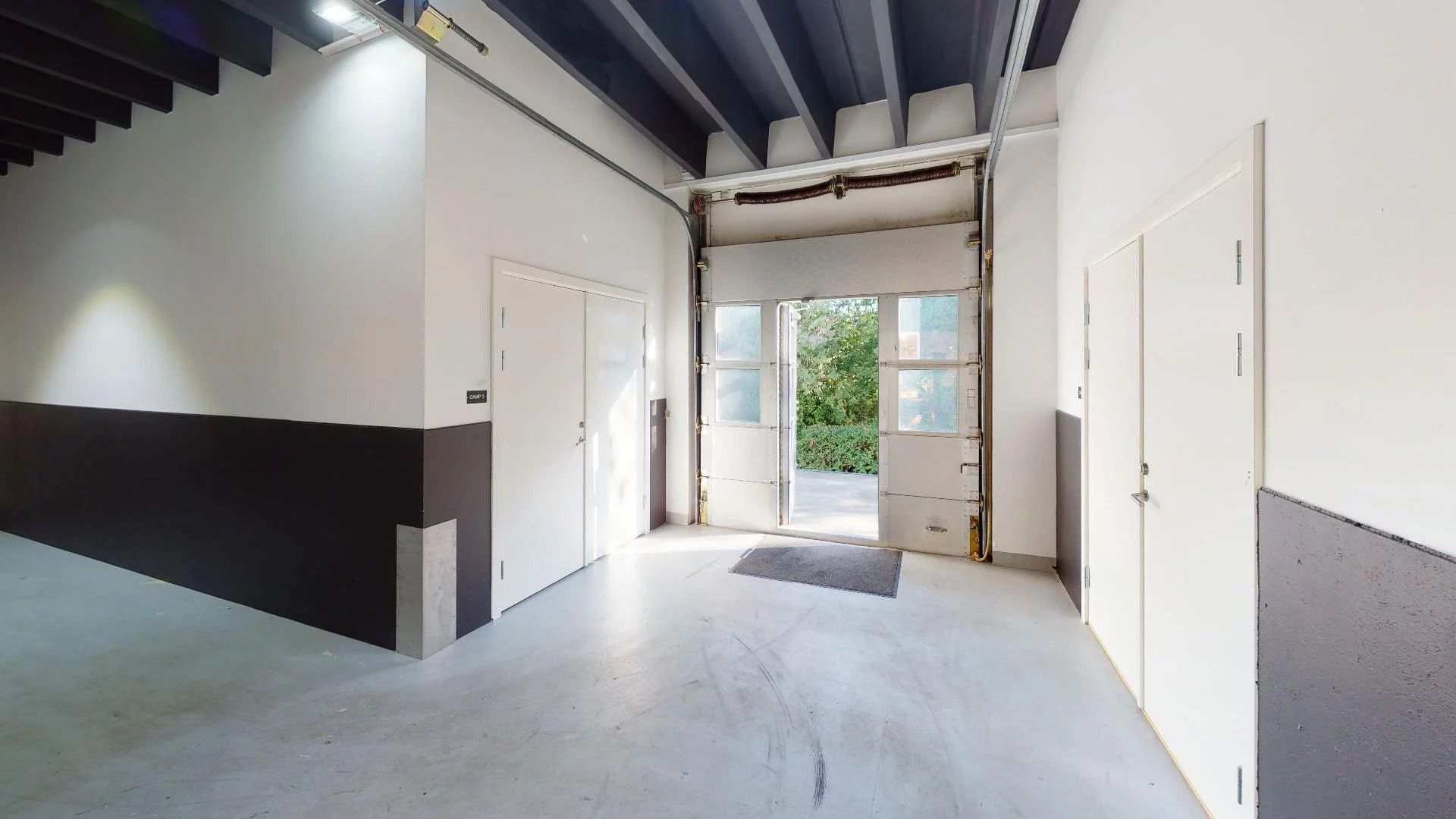 En lagerhal med hvide vægge og gulv samt højt loft med bjælker og to åbne døre og et glasvindue