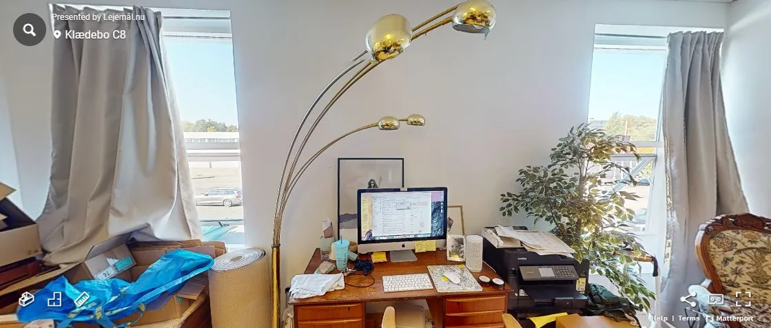 Kontor med et bord og computer samt en lampestang og en plante udenfor vinduet