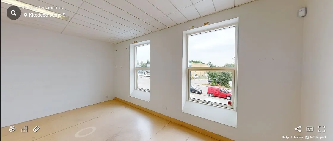 En tom stue med to store vinduer og en snedækket parkeringsplads udenfor med en rød bil og en sort bil i afstand