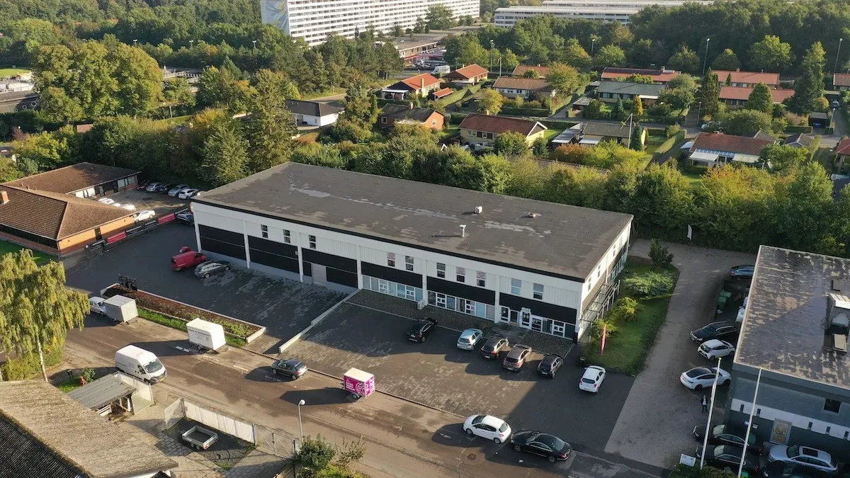 Firmabygning med stor parkeringsplads og adgangsvej i forgrunden og en bygning med røde tag i baggrunden