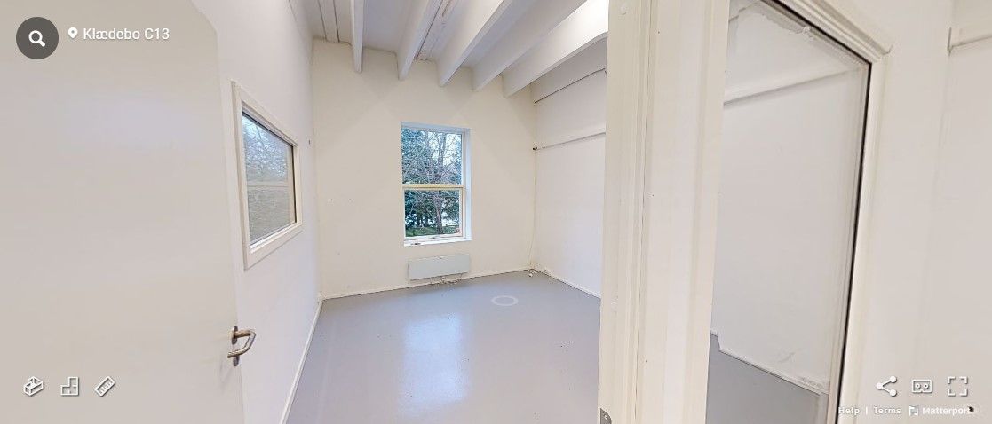 Et tomt værelse med hvide vægge, to vinduer og en hvid gulvbelægning, der vender mod en åben dør med håndtag