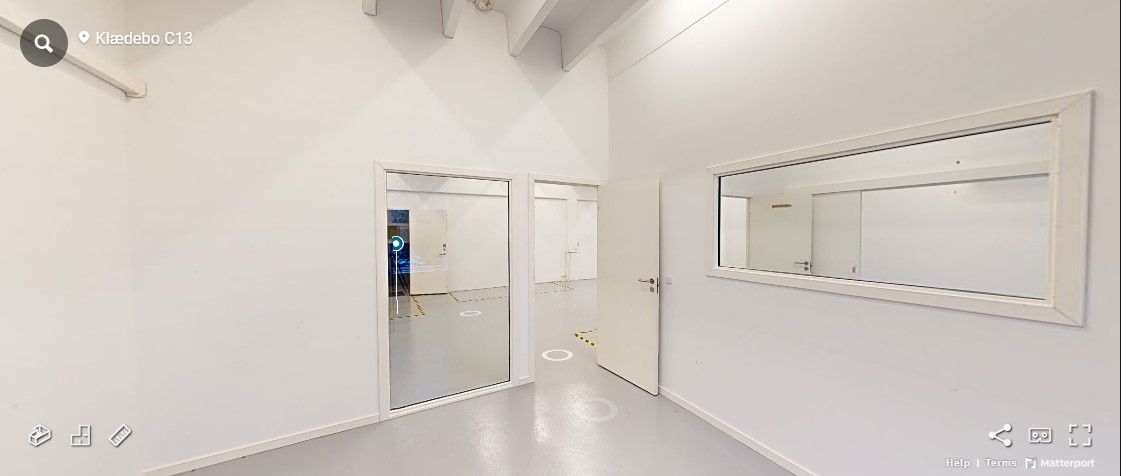 Rummelige lokaler med hvide vægge og glasdøre og spejl. Et mindre rum med lys og gul gulvmærkning. På væggen er der tekst,