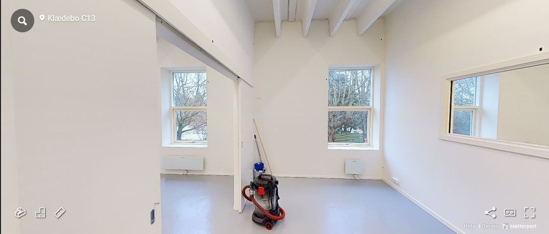 Et tomt kontorlokale med hvide vægge og gulve samt to vinduer med udsigt til træer og en støvsugemaskine i midten