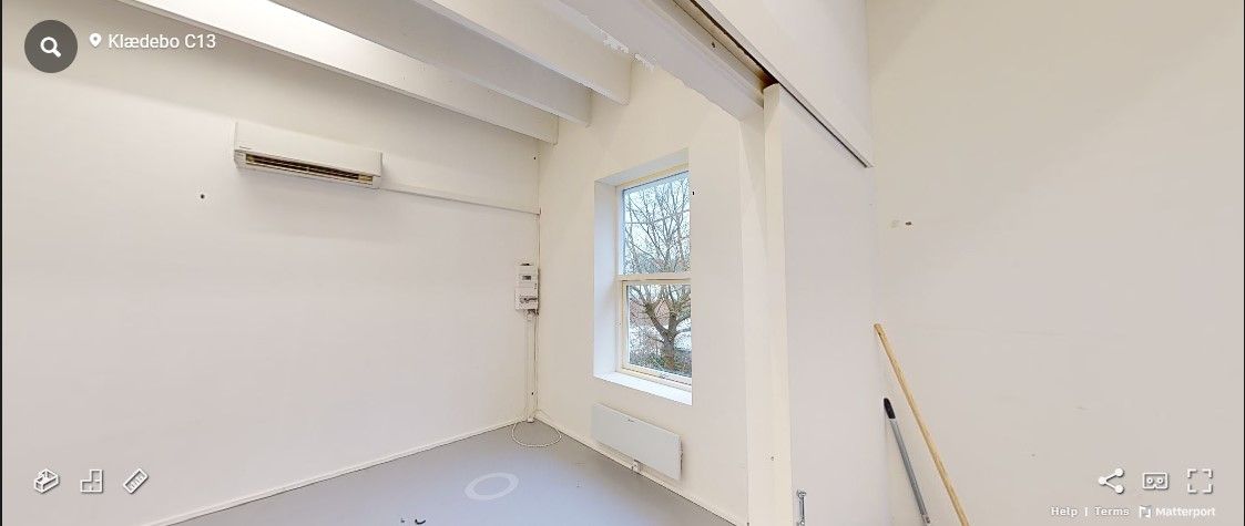 Værelse med hvide vægge, klimaanlæg, radiator og vinduesglas, hvor træerne er synlige udenfor. Rensevippe og udstyr står i