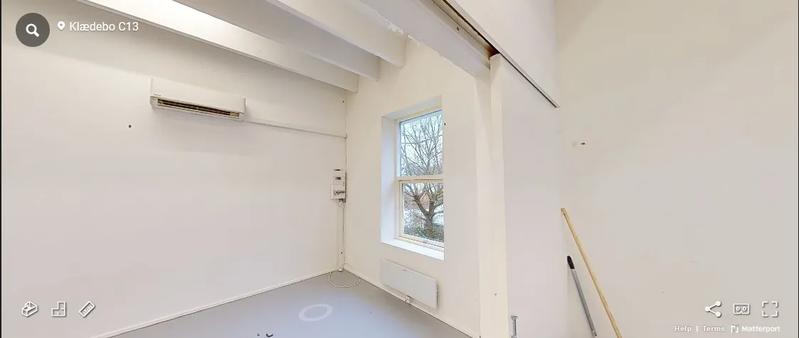 Værelse med hvide vægge, klimaanlæg, radiator og vinduesglas, hvor træerne er synlige udenfor. Rensevippe og udstyr står i