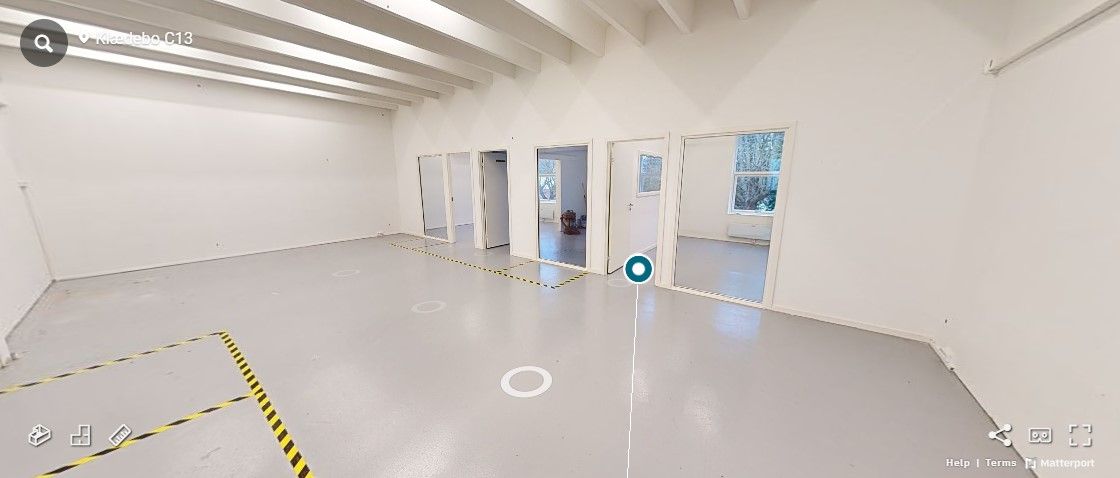 En stor, tom kontorlokal med hvide vægge, gulv og loft. I midten er der en stor, tom gulvplade. I baggrunden er der fem døre