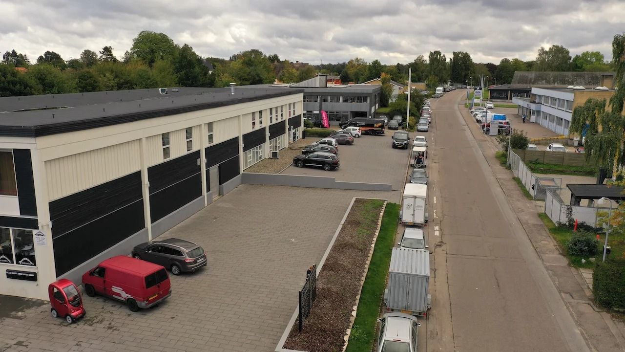 Biler og lastbiler parkeret i erhvervsejendom med facader i hvidt og sort og et banner med pink farve på en side af bygningen