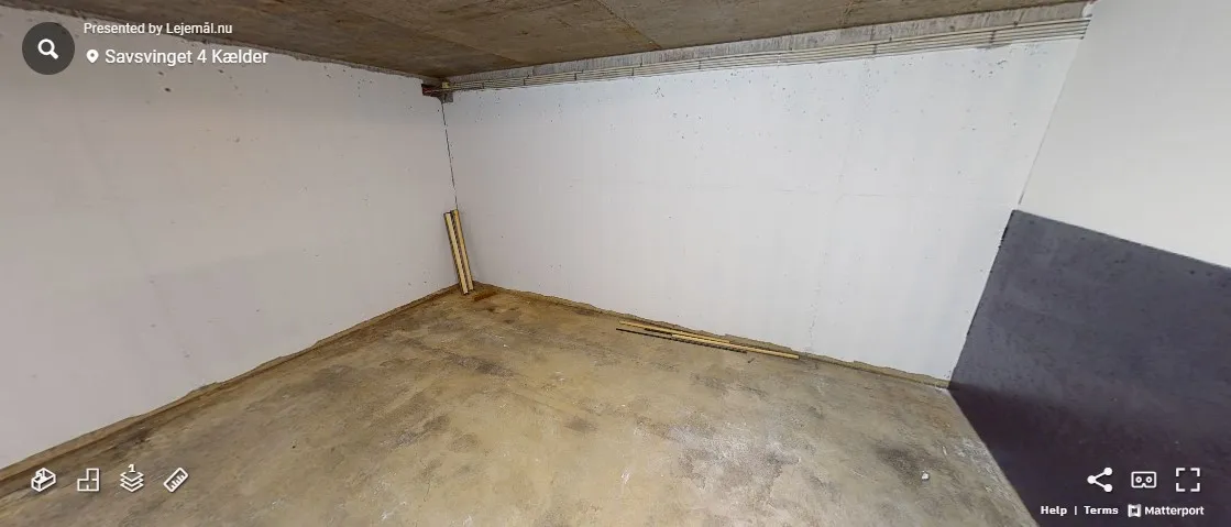 Lagerrummet er ubeboet med betongulv og hvide vægge. Et stykke træ ligger på gulvet. Der er plads til flere ting i rummet.