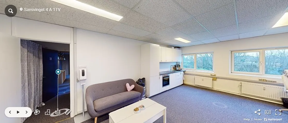 Inde i et lejlighedsværelse med sofa, kaffebord og køkken med ovn, vandhaner og vinduer mod gaden